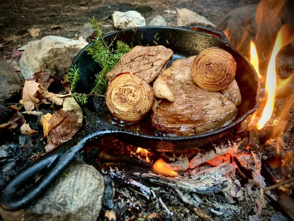 A steak fries in a cast iron pan over a camp fire