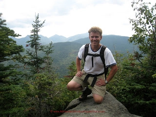 A man on a mountain top wearing a backpack smiles into the camera
