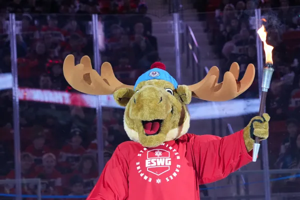 A moose mascot wearing a red Empire State Winter Games jersey holds a flaming torch aloft during an opening ceremony.