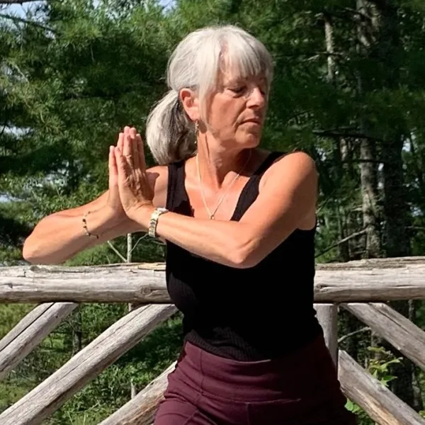 A woman holds a yoga pose with her eyes closed outside surrounded by pine trees.