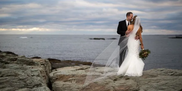 Bride and groom stand on a rocky surface overlooking a lake.