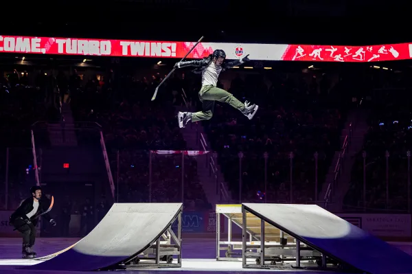 An ice skater performs a high jump over a ramp on an ice rink, holding a hockey stick as part of a stunt performance.
