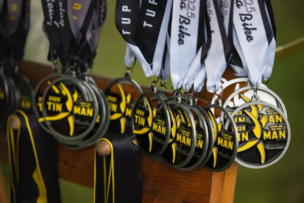 A row of silver 2025 Tupper Lake Tinman finisher medals with yellow and black accents hang from ribbons on a wooden rack.