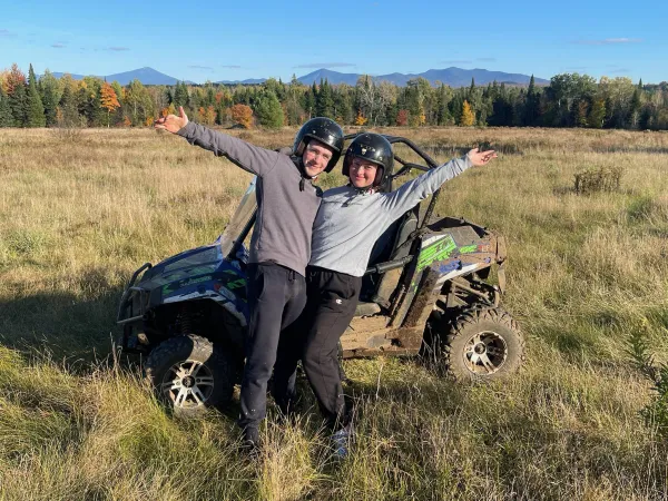 A happy couple smile into the camera with an arm raised in excitement in front of a UTV