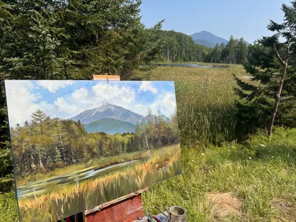 The artist's view of a plein air painting underway while still attached to an easel while still overlooking the field, pine trees, swamp surrounding a river and mountain view that inspired the art.