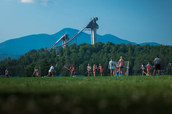 Female lacrosse players in white jerseys play against team in orange jerseys on field in front of mountains and ski jumps