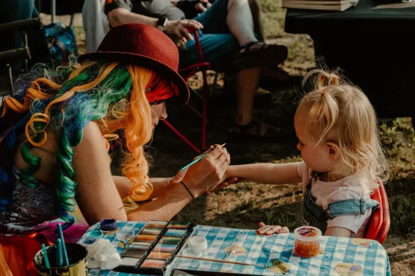 Woman wearing rainbow colored wig paints on the hand of a small child with art supplies on table in front of them
