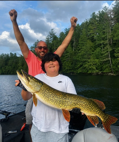 A kid holds a large pike while a man throws up his hands in victory behind.