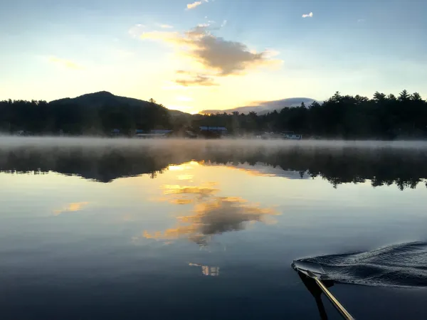 Mist rises over a perfectly still lake with mountains in the background.