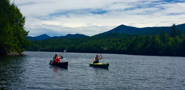 Two kayakers paddle down the water with mountains in the background.