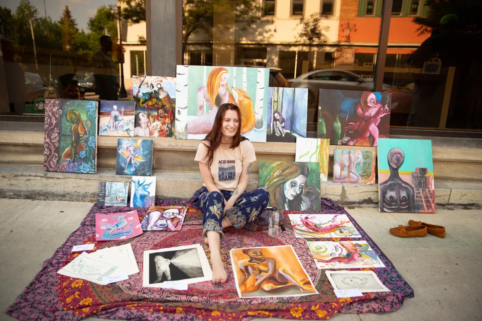 A woman sits proudly with her artwork on a sidewalk. 