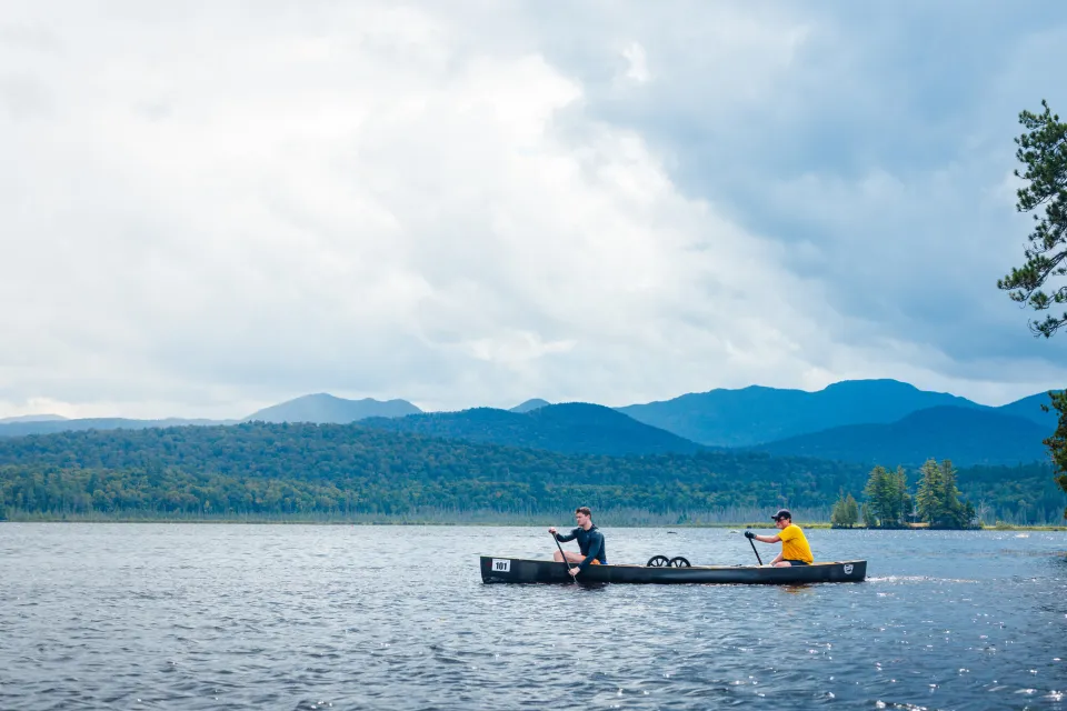 Two adults paddle a long canoe on a lake with mountains in the distance.