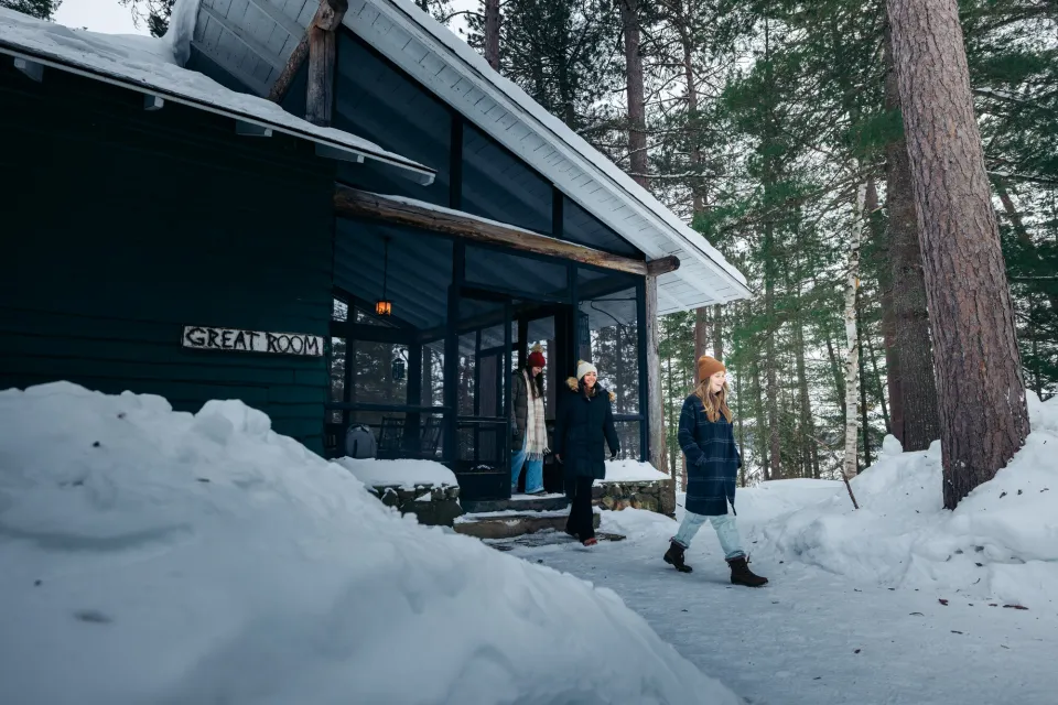 Three women walk in winter clothes out of a snowy cabin.