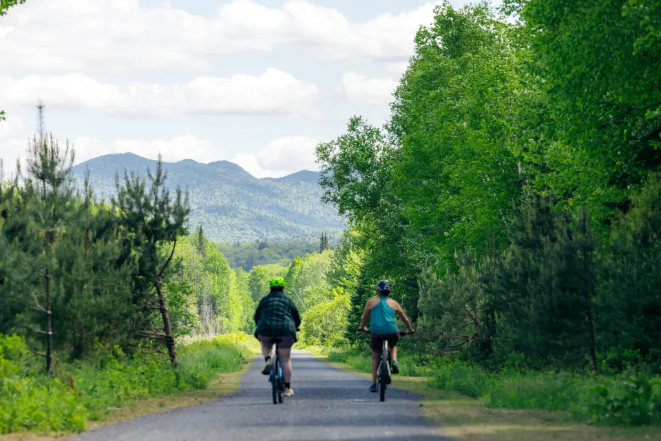Two women ride bicycles on a paved path with mountains in the distance.